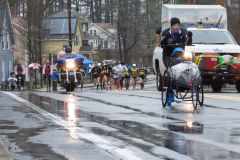 Photo Racer pushing a wheelchair as the first women (and their press and police trucks) come up behind them.