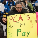 Woman with sign "PCAs need more pay" indoors