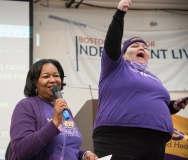 Two woman speak to the crowd, wearing purple 1199SEIU shirt , indoors