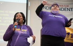Two woman speak to the crowd, wearing purple 1199SEIU shirt , indoors