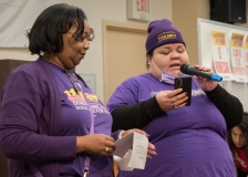 Two woman speak to the crowd, wearing purple 1199SEIU shirt , indoors