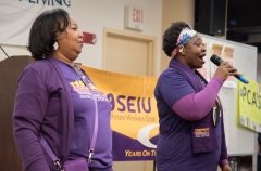 Two woman speak to the crowd, wearing purple 1199SEIU shirt , indoors