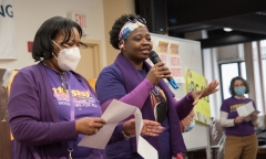 Two woman speak to the crowd, wearing purple 1199SEIU shirt , indoors