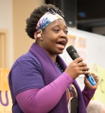 One woman speak to the crowd, wearing purple 1199SEIU shirt , indoors