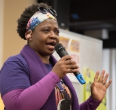 One woman speak to the crowd, wearing purple 1199SEIU shirt , indoors