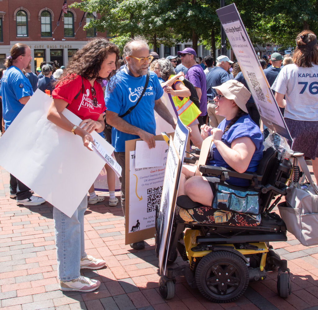 Kay (BCIL) and Rick Glassman speaking to woman in wheelchair with lots of signs