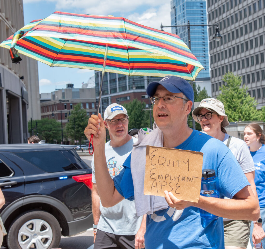 Man with shade umbrella and sign: Equity Employment APSE