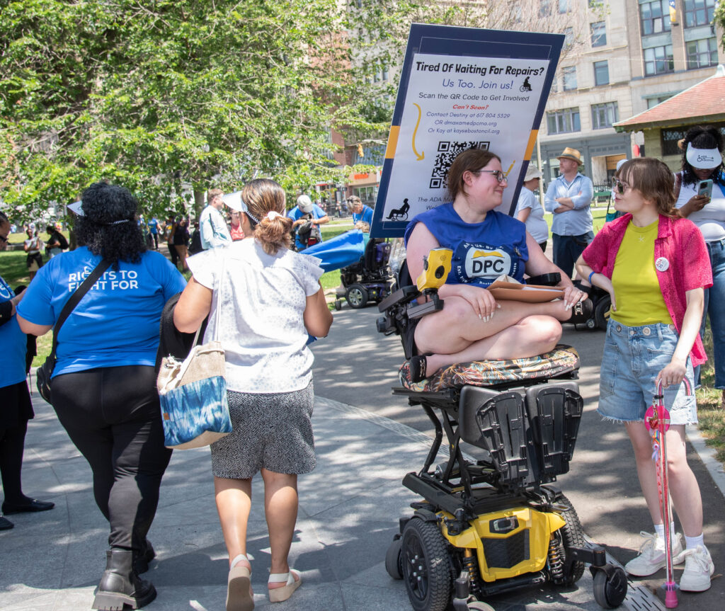 Women talking - one in wheelchair with Wheelchair repair protest sign