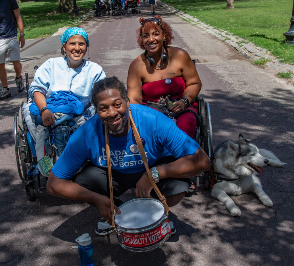 2 women in wheelchair, man squatting in front with a drum