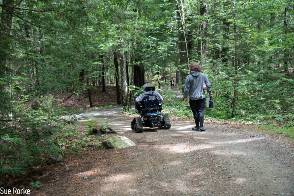Man in all terrain chair goes down wooded path with friend